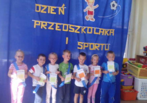 Grupka dzieci w niebieskich szarfach pozująca do zdjęcia trzymająca przed sobą dyplomy.