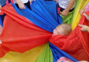 Dziewczynka zawinięta w chustę animacyjną przez inne dzieci podczas zabawy. Widoczna jest tylko jej uśmiechnięta buzia.