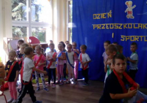 Dwa zespoły dzieci w szarfach podczas przygotowania konkurencji sportowej.