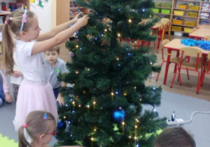 Dziewczynka zawiesza bombkę na choince, lśniącej światłem lampek.