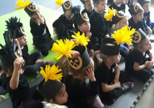 Na macie siedzą dzieci, na głowach mają czapki górnika czarne z żółtymi pióropuszami