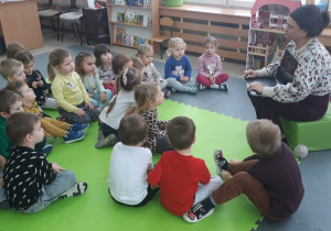 Nauczycielka siedzi na pufie i trzyma w dłoni komputer, dookoła niej siedzą dzieci