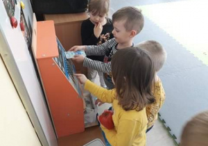 Dzieci uczą się korzystać z bankomatu. Przy drewnianym bankomacie stoi czworo dzieci