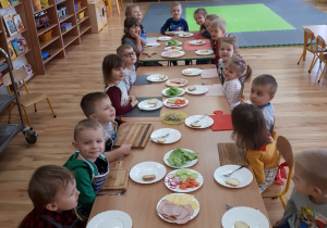 Dzieci siedzą przy połączonych ze sobą stolikach, na których ustawione są talerzyki z bułkami, a także talerze z produktami do kanapek.