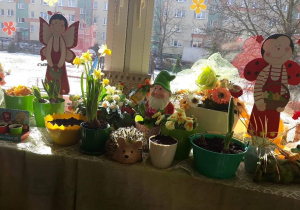 Na parapecie stoją doniczki z kwiatami, w tle dekoracja; krasnal i dwie biedronki