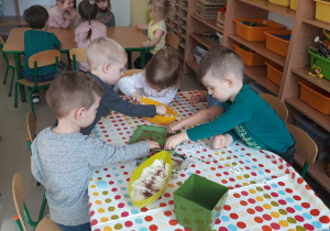 Na stole stoją pojemniki z nasionami, dzieci trzymają dłonie w pojemnikach