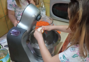 Dwie dziewczynki w kuchni. Jedna siedzi na blacie kuchennym, druga zaś stoi przy robocie kuchennym wbijając do niego jajka.