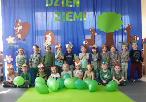 Dzieci pozują do wspólnego zdjęcia, na głowach mają ekologiczne opaski,w tle dekoracja z napisem "Dzień Ziemi"