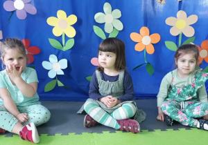 Trzy dziewczynki siedzą po turecku, w tle są kwiaty