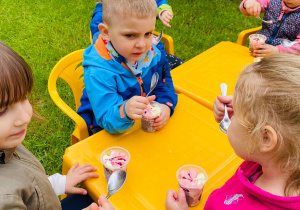 Grupa dzieci zjada lody siedząc przy stoliczkach.