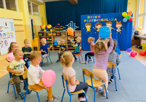 Dzieci siedzą na krzesełkach i podają sobie balony.