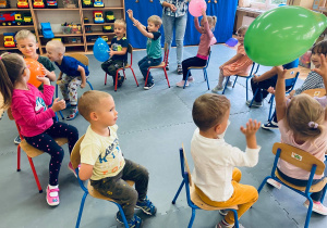 Przedszkolaki siedzą na krzesełkach i podają sobie baloniki.