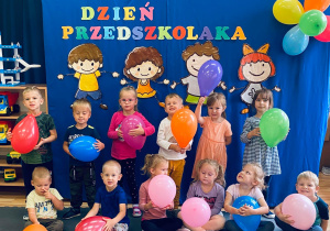 Przedszkolaki pozują do zdjęcia trzymając kolorowe balony na tle dekoracji.