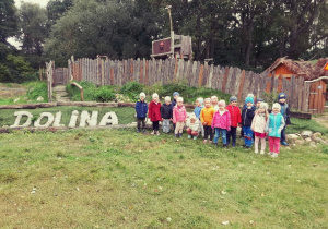 Dzieci pozuja do zdjęcia przed ogrodzeniem Doliny Skrzatow. W tle napis Dolina Skrzatow
