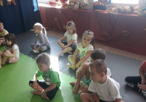 Dzieci siedząc w siadzie skrzyznym uważnie słuchają poleceń nauczycielki