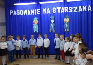 W głębi sali granatowa zasłona, na niej biały napis: "Pasowanie na Starszaka", dzieci ubrane na galowo stoją w półkolu