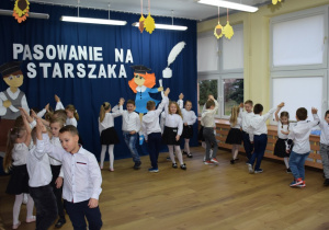 W głębi sali granatowa zasłona, na niej biały napis: "Pasowanie na Starszaka", dzieci ubrane na galowo tańczą w parach