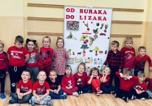 Grupa dzieci ubranych na czerwono pozuje do zdjęcia przy tablicy z ilustracjami i napisem Od buraka do lizaka.