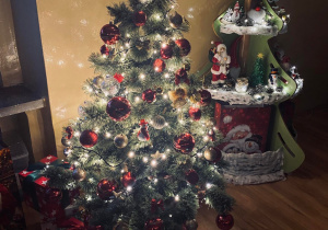 Choinka strojnisia pięknie ubrana przez przedszkolaki. Obok kącik z świąteczno - zimowymi dekoracjami.