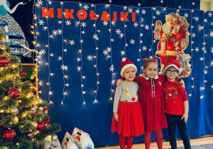 Jula, Tosia i Milenka pozują w Mikołajkowych czapkach do wspólnego zdjęcia na tle dekoracji.