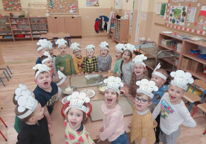 Przedszkolaki uśmiechając się stoją wokół złączonych stolików w opaskach - czapkach szefów kuchni.