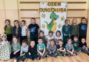 Grupa dzieci pozuje przy tablicy z napisem DZIEŃ DINOZAURA i zielonym dinozaurem.
