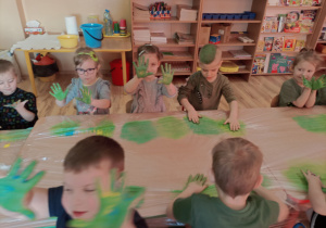 Dzieci przy stoliczkach pokazują rączki pomieszaniu zielonej farby.