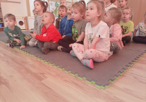 Przedszkolaki siedzą na macie i oglądają film edukacyjny o dinozaurach.