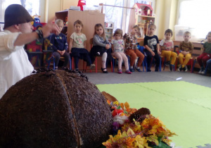 Na pierwszym planie imitacja mrowiska, w tle dzieci siedzące na krzesłach i słuchające opowieści prowadzącego.