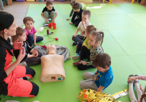 Dzieci siedzą przy fantomie, słuchają w jaki sposób można udzielić pierwszej pomocy