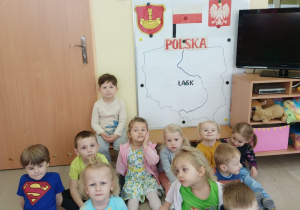 Dzieci siedzą przed tablicą z informacjami o Polsce i Łasku