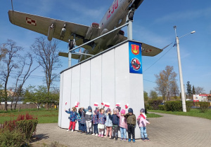 Dzieci stoją pod pomnikiem samolotu z flagami