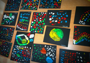 Zakropkowane prace przedszkolaków wykonane farbami na czarnym kartonie.