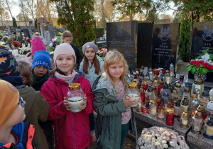 Dwie dziewczynki trzymające w rękach znicze, w tle dzieci przy grobach poległych żołnierzy