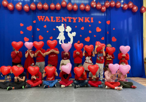 Dzieci ubrane na czerwono z walentynkowymi balonami w kształcie serc pozują do wspólnego zdjęcia na tle walentynkowej dekoracji.