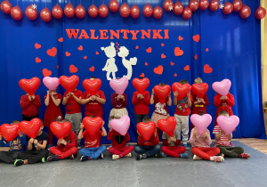 Dzieci ubrane na czerwono z walentynkowymi balonami w kształcie serc pozują do wspólnego zdjęcia na tle walentynkowej dekoracji.