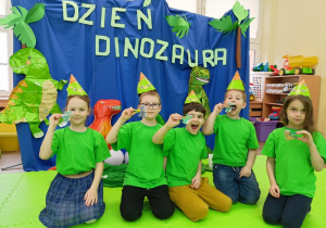 Grupka dzieci w czapkach dinozaurów pozuje do zdjęcia wraz z wykonanymi dinusiami na tle dekoracji.