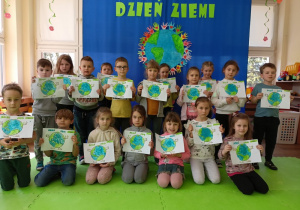 Cała grupa dzieci pozuje do zdjęcia ze swymi pracami na tle dekoracji z okazji Dnia Ziemi.