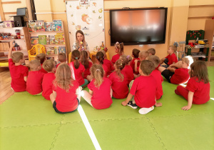 Dzieci w czerwonych koszulkach zwrócone są do nauczycielki, która trzyma w ręku jabłko.