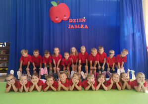 Ubrane w czerwone koszulki przedszkolaki pozują do zdjęcia pod napisem dzień jabłka.