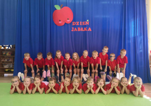 Ubrane w czerwone koszulki dzieci pozują do zdjęcia pod napisem dzień jabłka.