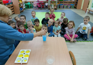 Dzieci siedząc na dywanie obserwują eksperyment z kolorami.